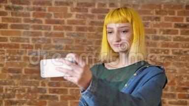年轻迷人的黄发女，带着手机自拍，微笑着站在砖墙背景附近，现代