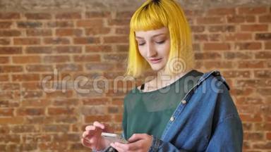 年轻不寻常的女人，一头<strong>黄发</strong>，拿着电话，看着摄像机，微笑着站在砖墙附近
