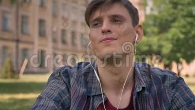 年轻的帅哥坐在公园的草地上，用耳机听音乐，唱歌，放松概念，<strong>继续前进</strong>