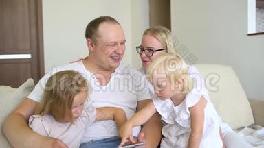 快乐的家庭一起玩笔记本电脑和数字平板电脑在家里。 爸爸，妈妈和两个孩子坐在沙发上