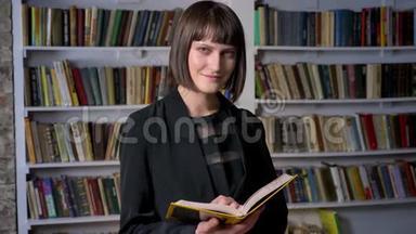 年轻漂亮的女人穿着夹克，拿着书在图书馆里看着相机，微笑着