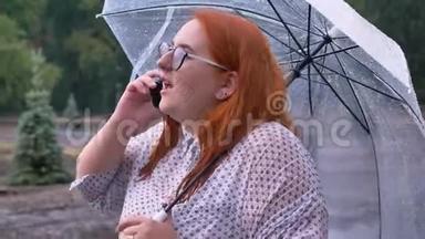 带眼镜的胖姜女站在公园的雨下，打电话，撑伞，交流