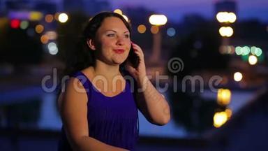 一位穿着蓝色连衣裙的年轻女子正在智能手机上交谈，背景是路边模糊的路灯。
