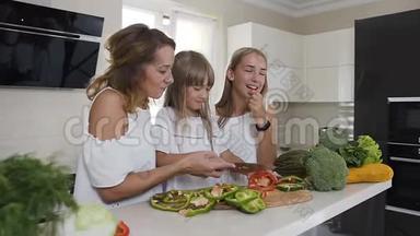 快乐的母亲和她的两个女儿正在家厨房做饭吃蔬菜。 食物，健康饮食，家庭