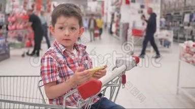 小男孩在智能手机上玩游戏，坐在手推车里，在超市的家庭购物中