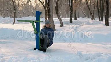 穿着带罩的蓝羽绒服的人正在冬季公园的模拟器上做腿部运动。 前景。