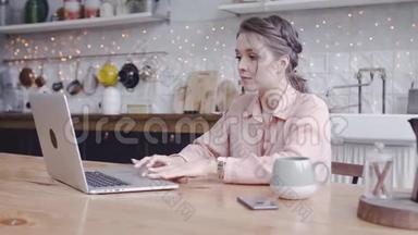 年轻漂亮的女人在家里厨房里用笔记本电脑工作。 <strong>库存</strong>录像。 漂亮的女人小心地在笔记本电脑上工作