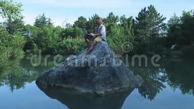 一个年轻的自由<strong>职业</strong>者坐在湖中央的石头上，手提电脑。 自由<strong>职业</strong>概念。 出差工作。 无线无线