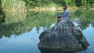 一个年轻的自由职业者坐在湖中央的石头上，手提电脑。 自由职业概念。 出差工作。 无线无线