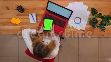 一位年轻的高加索员工使用平板电脑的特写镜头，平板电脑的绿色屏幕坐在室内笔记本电脑前