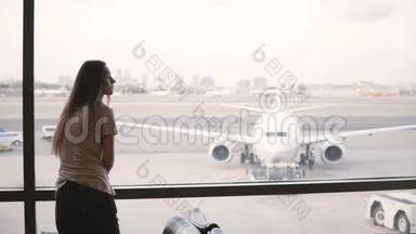美丽的女游客站在机场候机楼的窗口看飞机，然后拿着包离开。