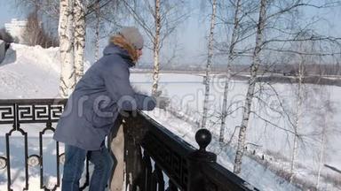 穿着蓝色羽绒服，带着毛皮帽的人站在栅栏旁，在冬季公园里环顾四周。
