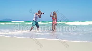 男摄影师拍摄年轻女子比基尼在海滩上摆姿势。 年轻的摄影师
