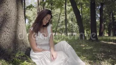 可爱的年轻女孩的肖像，长着一头深色长发，穿着一件白色的夏季时装长裙，<strong>坐在树下</strong>。