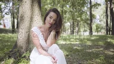 可爱的年轻女孩的肖像，长着一头深色长发，穿着一件白色的<strong>夏季</strong>时装<strong>长裙</strong>，坐在树下。