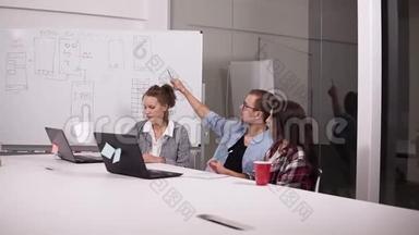 戴眼镜的年轻人和两个随意坐在办公室桌子上的女人在创意工作场所，上面有笔记本电脑。 男子