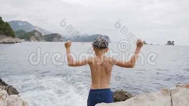 后景。 可爱的孩子举起双手站在海边的岩石上。 快乐的男孩高<strong>高举</strong>起双手