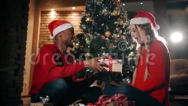 幸福的一对夫妇正坐在家里客厅的一块蓬松的地毯上。 一个男人正在给女孩送圣诞礼物。