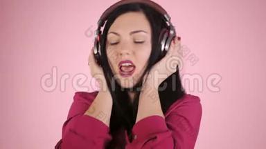 戴着耳机听着音乐的美女，闭着眼睛站在衬衫上的粉色背景上