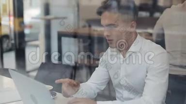 快乐的年轻商人坐在一张桌子上，在一家咖啡馆里用笔记本电脑工作。 赢家看手提电脑显示是的手势