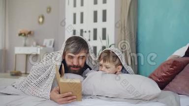 可爱的父亲和可爱的儿子正在读可怕的故事，躲在毯子下，躺在家里的床上。 快乐