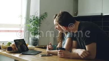 年轻漂亮的白种<strong>人</strong>夫妇<strong>坐</strong>在现代化的厨房里，<strong>坐</strong>在笔记本<strong>电脑前</strong>的桌子上，两个女孩在空中飞舞，他们是。