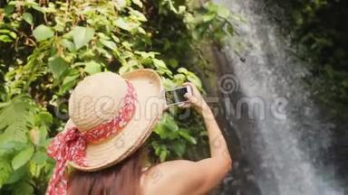 年轻的快乐旅游女孩穿着白色连衣裙和草帽<strong>制作</strong>照片惊人的丛林瀑布使用移动智能<strong>手机</strong>。 自然