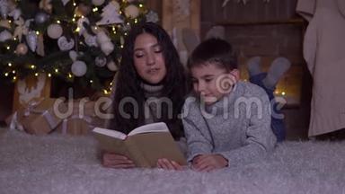 留着长发的妈妈给儿子读一本书，并在圣诞节的时候亲吻他躺在圣诞树附近的地板上。 <strong>高清高清</strong>