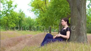 一个年轻漂亮怀孕的女孩穿着一件黑色T恤<strong>坐在树下</strong>