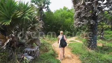 女孩游客带着背包，沿着热带岛屿森林的小径行走。 手掌和大海