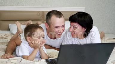 幸福的家庭：爸爸，妈妈和宝宝，躺在床上，看着笔记本电脑，慢悠悠地笑着..