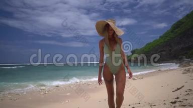 穿着<strong>绿</strong>松石比基尼和戴着一顶<strong>大</strong>草帽的<strong>漂亮</strong>成年女孩在热带海滩的沙滩上散步，背景是
