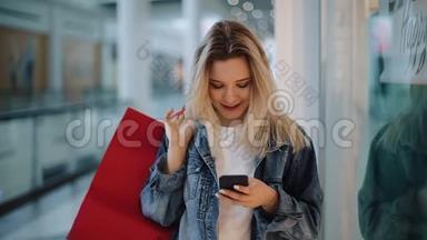 微笑的金发女孩在手机里输入一些东西，带着购物袋在购物中心周围<strong>走动</strong>