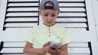 少年戴着帽子在智能手机上玩游戏，孤立在白色的门背景上。 儿童玩智能手机