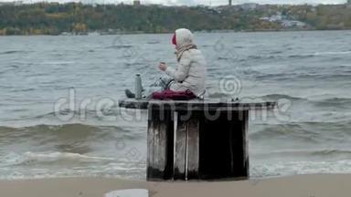 年轻女子穿着暖和的衣服坐在海边，坐在木盘管上，用热水瓶喝热茶