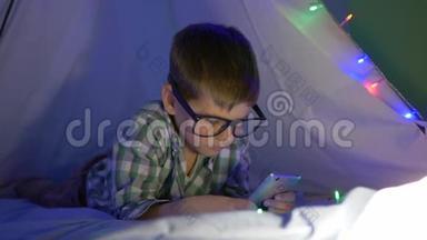 手机小孩，戴眼镜的小男孩正在玩智能手机，躺在wigwam上戴着花环