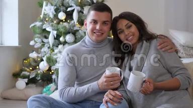 早上，在圣诞树的背景下，丈夫和妻子手里拿着白色的大杯子在家里的沙发上