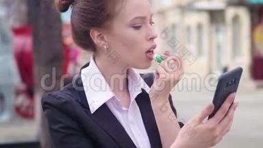 街上的商务女士用一台面向前方的智能手机相机作为镜子来描绘嘴唇。