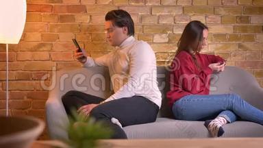 年轻的白种人夫妇背靠背坐在沙发上，在家庭氛围中使用智能手机的肖像。
