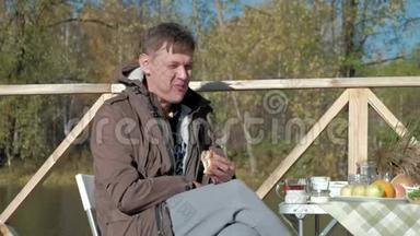 成熟的男人，穿着温暖的衣服，坐在桌子旁，吃着汉堡包，微笑着，从热水瓶里倒茶，野餐