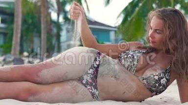 穿比基尼的年轻女人躺在海边，把沙子撒在你的身上。 有魅力的女人在沙滩上玩耍