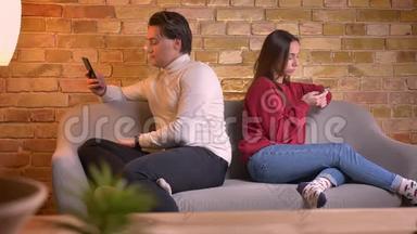 年轻的白种人男女背<strong>靠背</strong>坐在<strong>沙发</strong>上，在家庭氛围中使用智能手机的肖像。