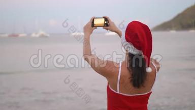 <strong>日</strong>出时，身穿圣诞帽和红色泳衣的女士在海滨用智能<strong>手机手机</strong>拍照