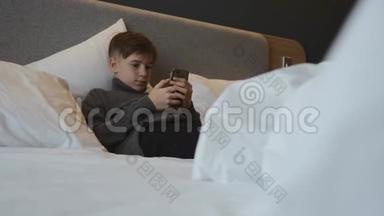 这个男孩躺在床上，手里拿着一部智能手机。 智能手机上的交流和游戏