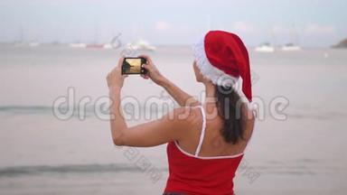 日出时，身穿圣诞帽和红色泳衣的女士在海滨用智能手机手机拍照