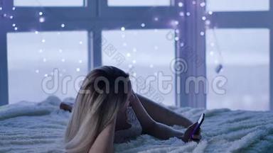 迷人的金发女郎在她的智能手机上写信息，躺在床上。 卧室里拿着手机的年轻女子