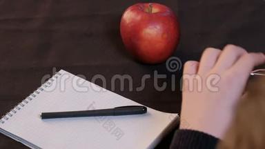 特写镜头。 一个人从桌子上举起眼镜。 一个红苹果躺在记事本和黑色<strong>布</strong>上的钢笔旁边。