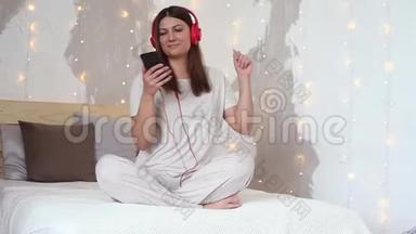 美丽的女孩听音乐耳机红色坐在床上，放松使用手机。 高清高清