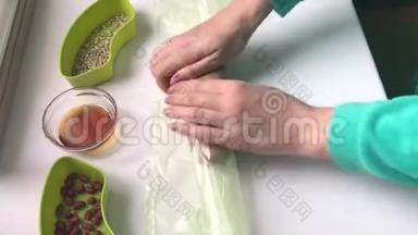 一个女人把一根香肠和碎花生、葵花籽和蜂蜜混合在保鲜膜里。 自制哈尔瓦烹饪。