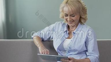 沙发上的老年妇女滚动着平板电脑屏幕，微笑着看着照片